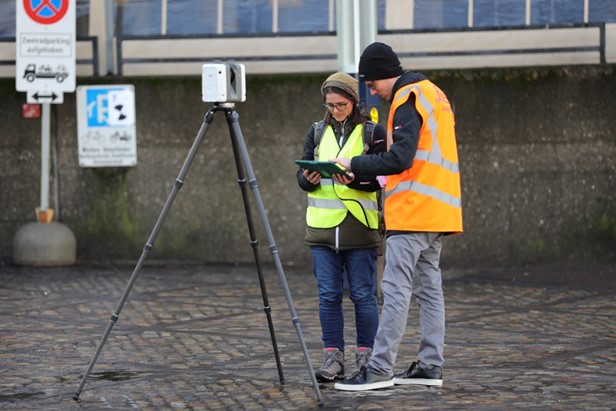 Zwei Personen in Signalwesten und mit einem Messgerät stehen auf einer Strasse und protokollieren Messergebnisse