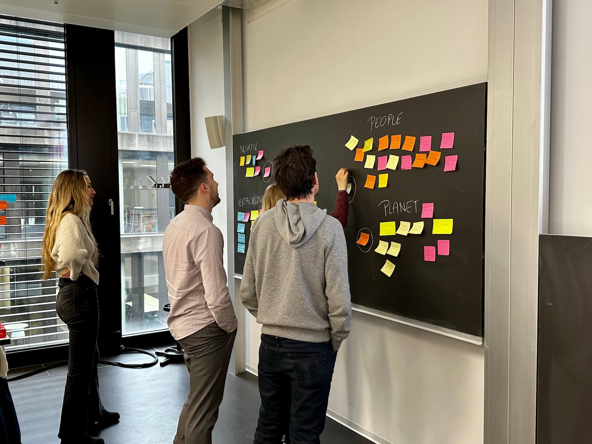 Studierende stehen vor einer Tafel auf die veschiedenfarbige PostIts im Rahmen einer Design-Thinking-Veranstaltung geklebt wurden