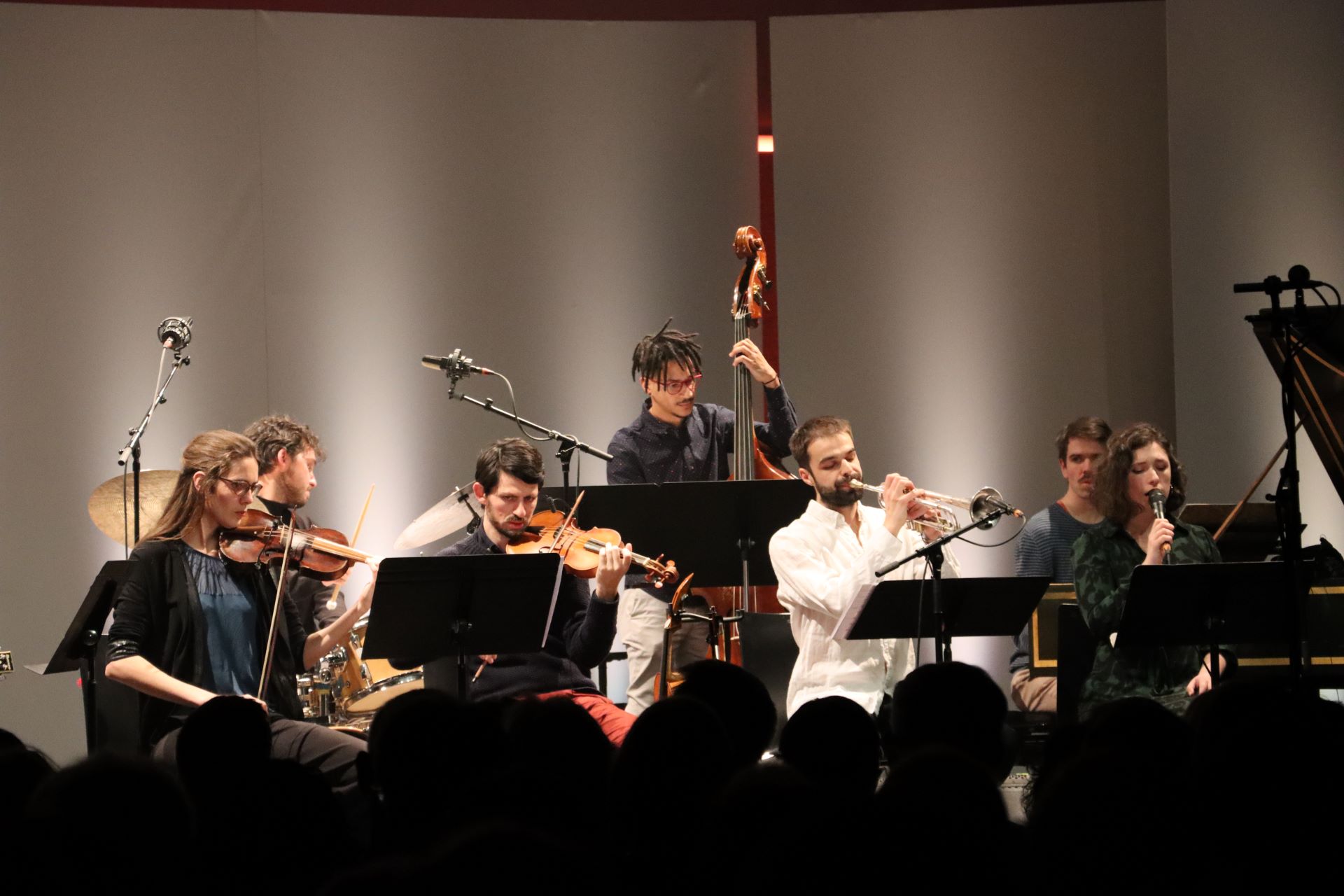 Mehrere Personen mit Instrumenten (Oboe, Geigen und Cello) stehen bzw. sitzen auf einer Bühne und musizieren. 