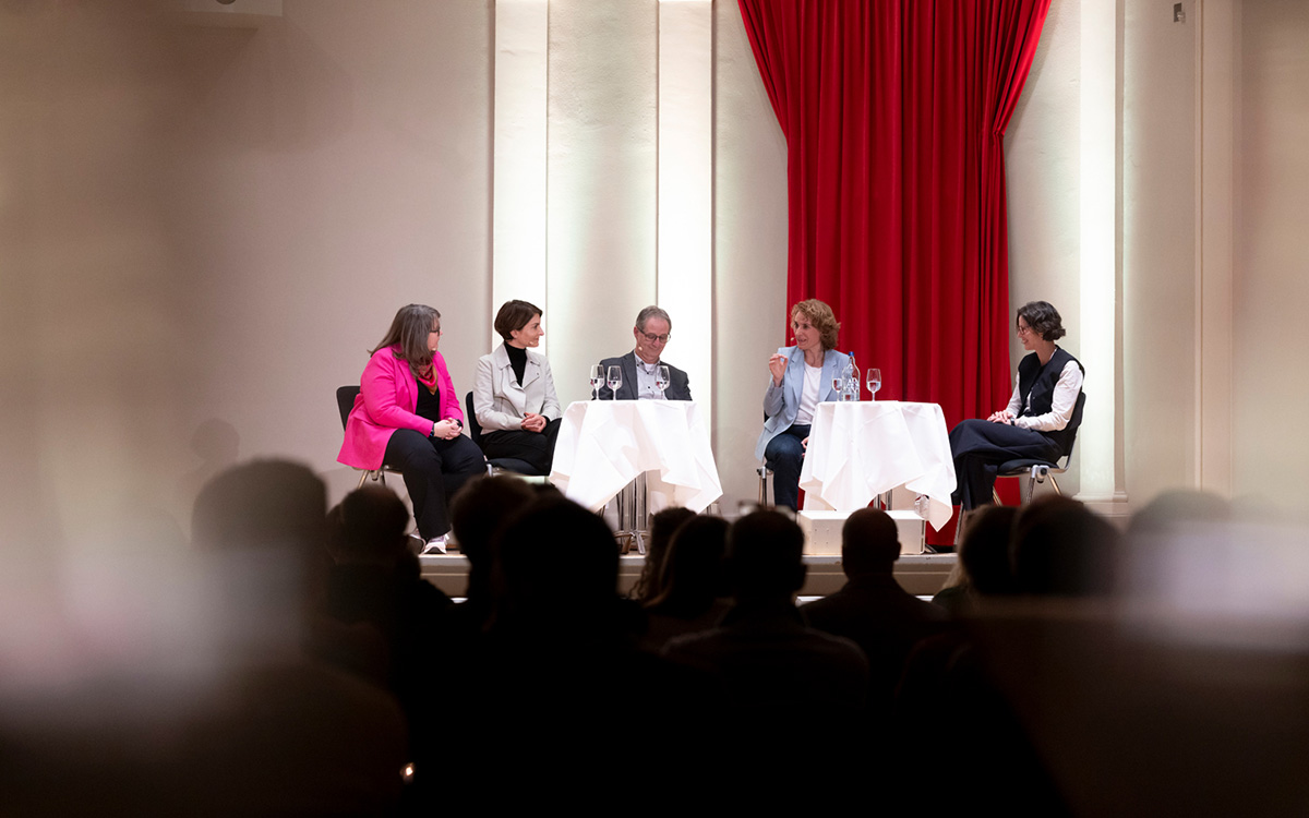 Fünf Personen sitzen auf der Bühne, an kleinen Tischen mit Mikrofonen und diskutieren. 