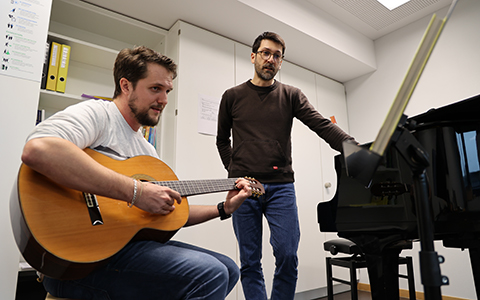 Gesangslehrer steht bei Student, der sitzend Gitarre spielt