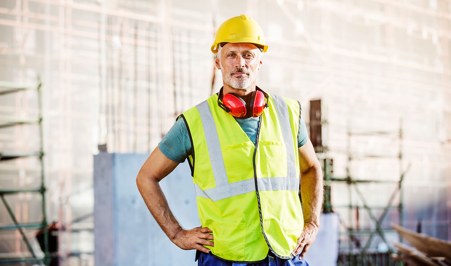 Ein Bauarbeiter steht vor einer Baustelle und schaut selbstbewusst in die Kamera.