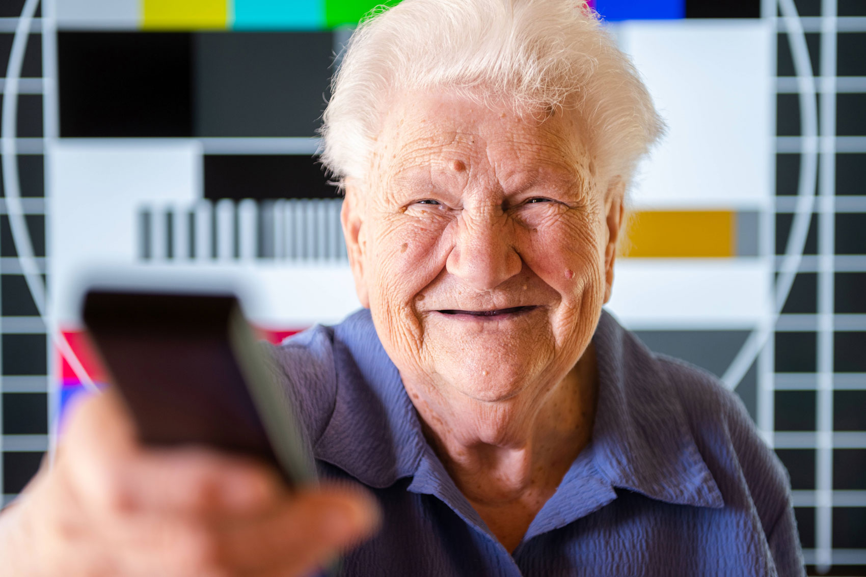 Ältere Frau hält lächelnd eine Fernbedienung in Richtung der Kamera