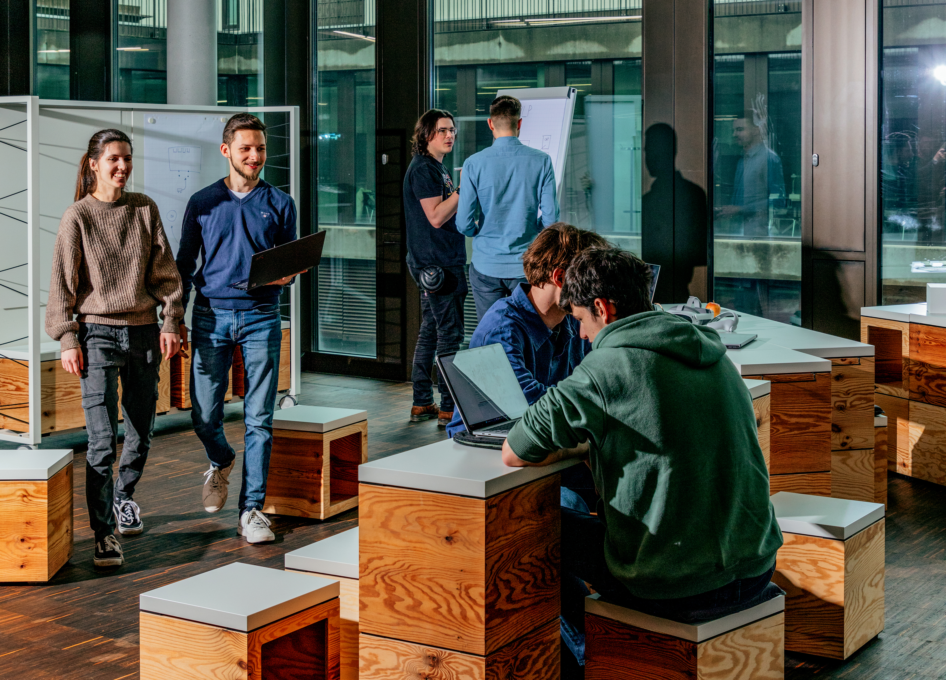Sechs Studierende stehen und sitzen zusammen an ihren Laptops und notieren Dinge auf einem Flipchart.