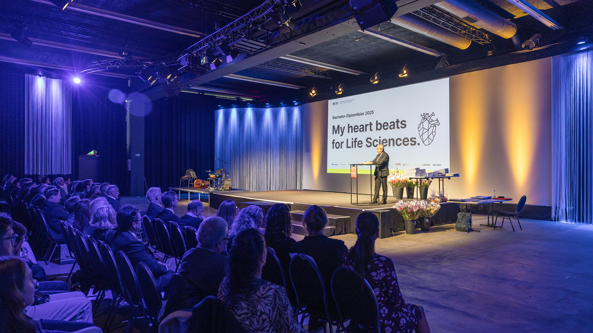 Ein Saal mit Bühne, im Hintergrund ist die Folie "My heart beats for Life Sciences" eingeblendet, ein Mann steht auf der Bühne an einem Rednerpult. Im Vordergrund sitzen Zuschauer*innen auf Stühlen. 