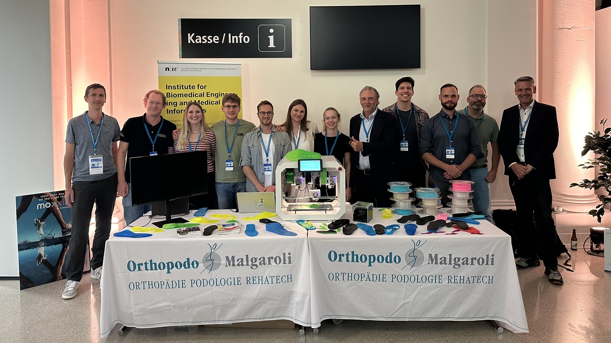 12 Personen stehen hinter einem Messetisch voller farbiger Sohlen aus dem 3D Drucker. 