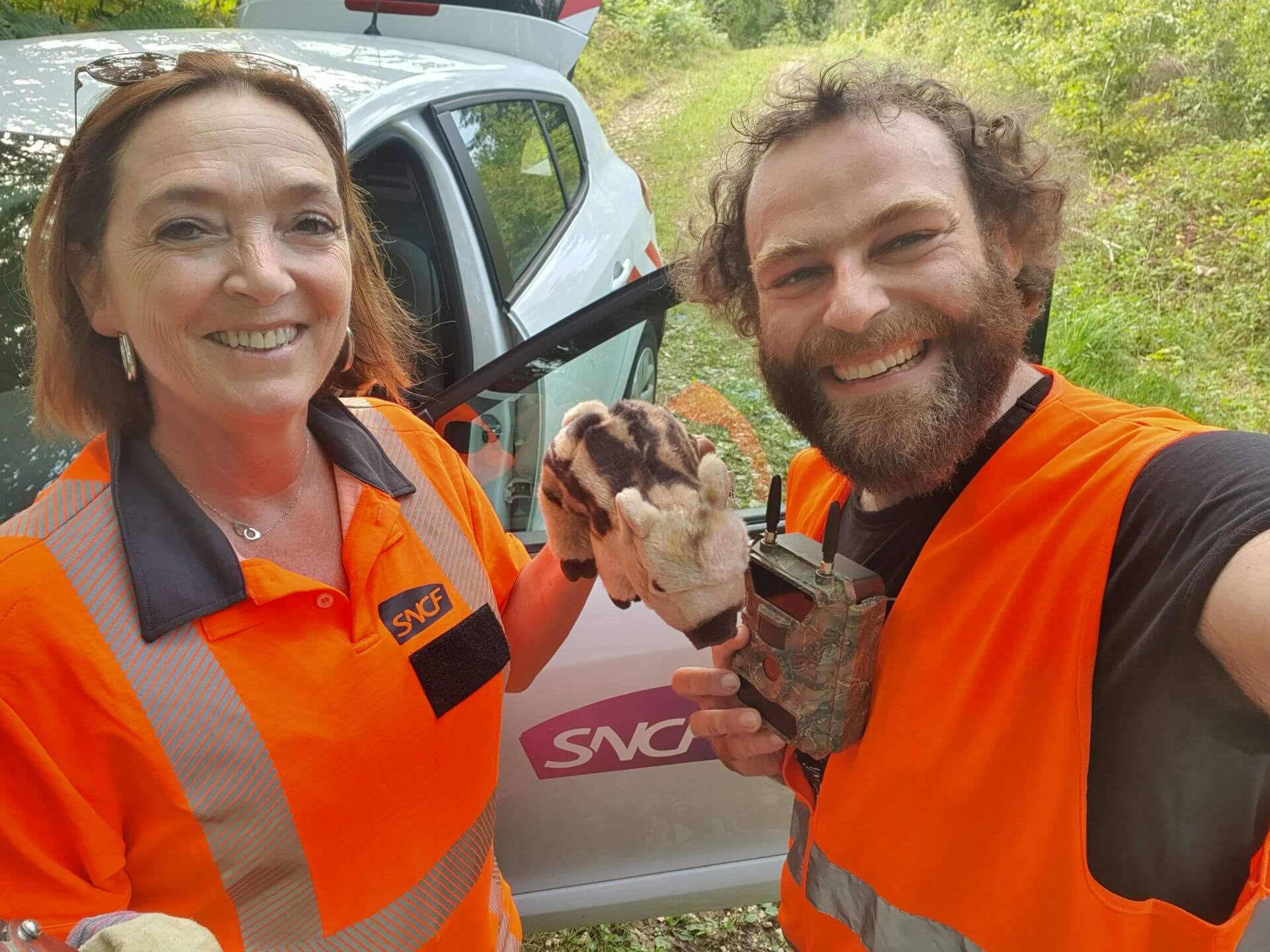 Selfie von Sylvie Perna und Adrian Meyer. Beide haben orangene Warnwesten an. Sylvie Perna hält ein Wildschwein-Stofftier in der Hand. Adrian Meyer ein Mesgerät