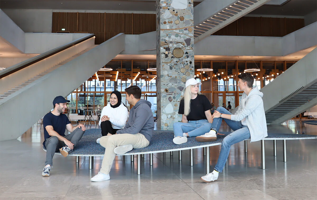 Fünf Personen sitzen im Eingangsbereich des FHNW Campus Muttenz und tragen die FHNW Merch-Kollektion. 