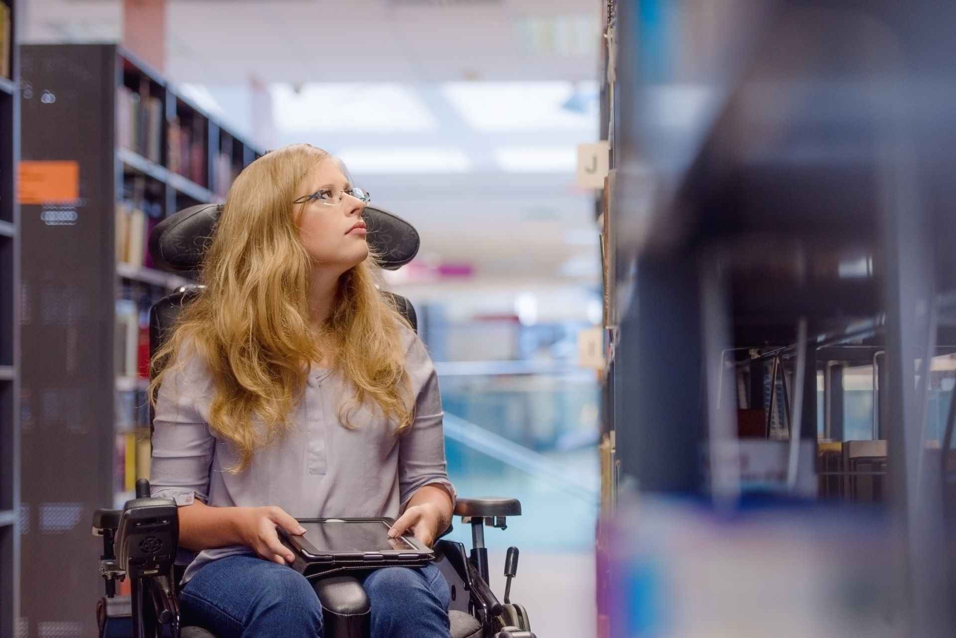A woman in a wheelchair holds a tablet in her hand, stands between two library walls and looks to the side. 